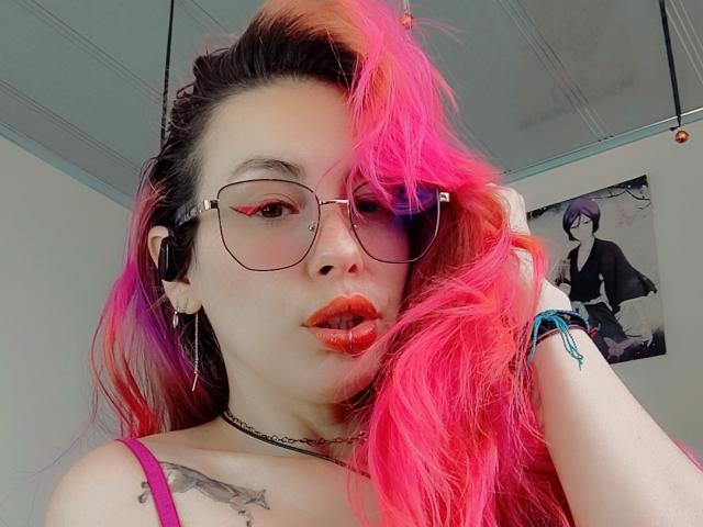 NatashaLau, with pink and purple dyed hair, wears glasses with black frames and red eyeliner. She has fair skin, red lipstick, and a black choker. Her left hand touches her hair, showing a tattoo on her right shoulder. She wears a pink strap top. The background includes a wall with a poster of a character in a black outfit. Her ear is pierced with a black earring. The image is a close-up selfie.
