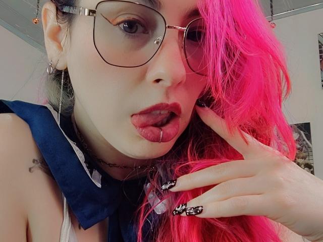 Close-up of NatashaLau with bright pink hair, wearing oversized glasses and a blue scarf. She has a septum ring in her nose and a lip ring. Her tongue is slightly out, and she's touching her cheek with a hand that has black and white manicured nails. She has a small tattoo on her upper left arm. The image focuses on her face and hair, with soft lighting highlighting her features. Her expression is slightly provocative.