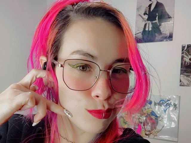 NatashaLau with pink and orange hair, glasses, red lipstick, and white nail polish, touching her ear, showing off her tits and ass with a cute smile.