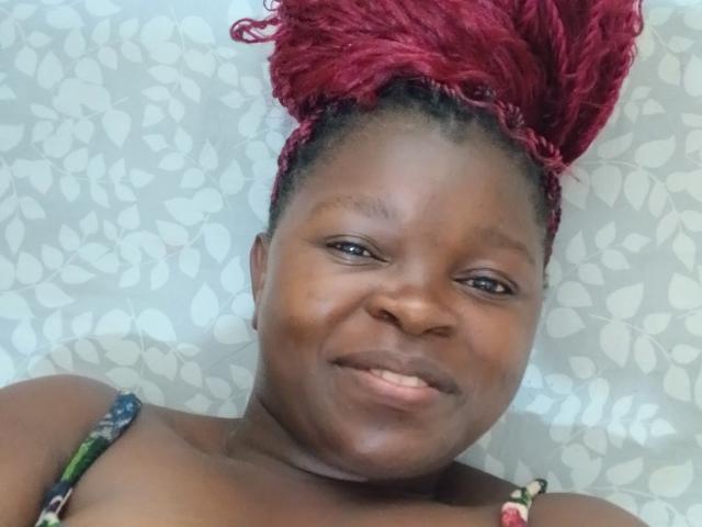 ZoeValerie with dark brown skin, red braided hair in a high bun, smiling slightly. She's topless with a colorful strap visible on her shoulder. Her face is centered, eyes looking sideways, and lips forming a subtle smile. The pattern on the background is white leaf-like shapes. The image is a close-up selfie. ZoeValerie's hair texture is tight and curly, contrasting with her smooth skin. The lighting is even, highlighting her facial features.