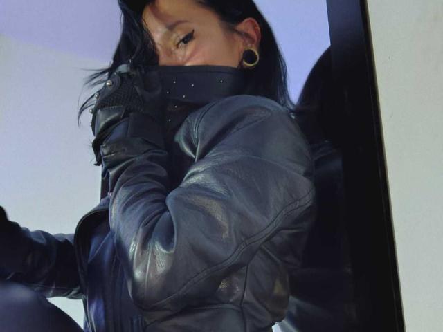 AmelieBlake is a woman with dark, slightly wavy hair, partially visible due to her black mask. She wears a black leather jacket with visible texture and a gold hoop earring in her left ear. Her right arm is bent, and her hand is near her face. The jacket's shiny, slightly worn texture contrasts with the matte black mask. The image is a close-up, focusing on her upper body and face. The lighting is soft, with a blueish tint.