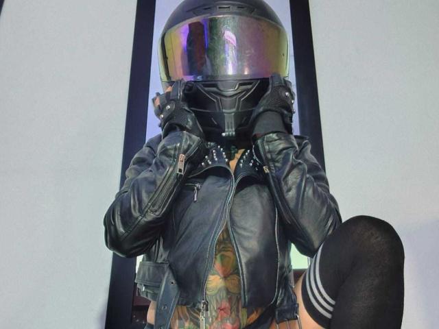 AmelieBlake wears a black leather jacket with studded shoulders and a shiny, reflective helmet. Her hands grip the helmet's sides. She has a colorful tattoo on her chest and a black knee-high sock on her left leg. The image focuses on her upper body and head. The jacket has a zipped front and a textured, worn look. AmelieBlake's face is obscured by the helmet. The photograph uses bright, even lighting.