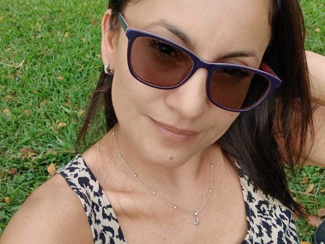 Close-up photo of SolaraDubois with medium skin tone, dark brown hair in a straight style, wearing blue-tinted sunglasses and a black-and-white leopard print tank top. She's smiling slightly, with a small mole on her right cheek. She's wearing a delicate silver necklace with a small cross pendant and small stud earrings. The grassy background is slightly out of focus. Her right arm is raised, partially visible. The overall composition is centered on her face and upper torso.