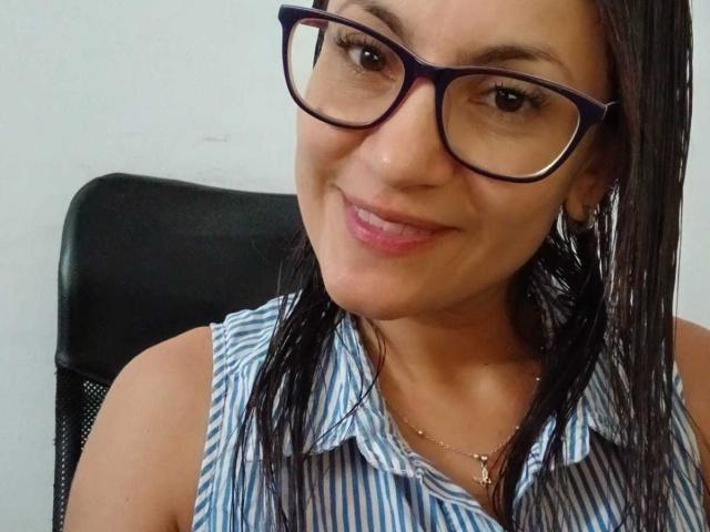 SolaraDubois with blue glasses, smiling, showing teeth, pink lipstick, and a small bit of her lower lip piercing visible, has brown eyes and dark hair, wearing a silver necklace.