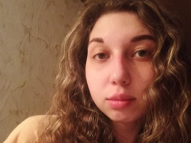 Close-up of Sanella with wavy, brown hair and fair skin. She has light brown eyes, subtle eyebrows, and pink lips. Her expression is neutral, slightly curious. The image focuses on her face, with light and shadow highlighting her facial features. The texture of her hair is curly and slightly messy. The background is a plain, beige wall with a subtle, patterned texture. Sanella's shoulders are partially visible, suggesting she is nude or wearing a strapless top. The image is casual and unfiltered.