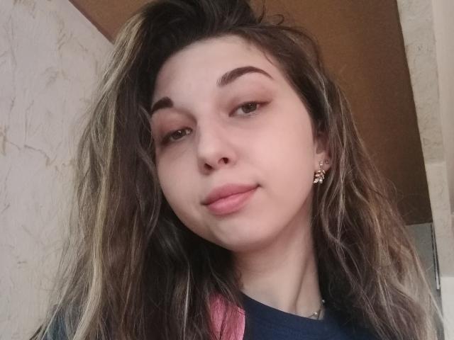 Sanella, a young woman with light skin and brown wavy hair, has a relaxed, slightly smug expression. She has thick, dark eyebrows and subtle pink eyeshadow. Her lips are full and pink. She wears a small gold earring in her left ear. Her hair is parted in the middle, and she has a small visible nose piercing. She wears a dark blue shirt with a pink pattern on the shoulder. The image is a close-up selfie.