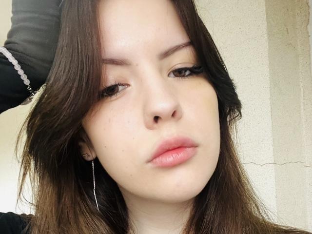 LynnEleanor's face with brown hair, slightly wavy, wearing black, has pink lips, small nose, light skin, and black eyeliner, holding a black garment, showing a neutral expression.