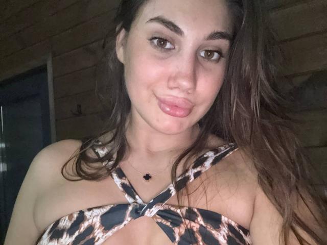 ErikaMelody with pouty lips, brown eyes, and long brown hair, wearing a leopard-print top, exposing her cleavage, and a black cross necklace, in a wooden room.