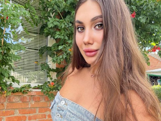 ErikaMelody has long, straight brown hair, light brown skin, and large brown eyes. She wears an off-the-shoulder denim top, exposing her collarbones. Her lips are slightly parted, showing a subtle pink. She has a small, circular necklace. The background includes a brick wall, green leaves, and red flowers. The composition focuses on her face, which is centered, with the greenery on the left. The image is bright and clear.
