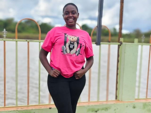 DestinyHot6_9 stands with hands in pink shirt pockets, smiling. She's a dark-skinned woman with a short, neat haircut. She wears a bright pink T-shirt with a cartoon teddy bear graphic and black pants. The background has a green and orange metal fence, a cloudy sky, and a river. Her stance is relaxed, and she looks confident. The image has a casual, everyday feel.