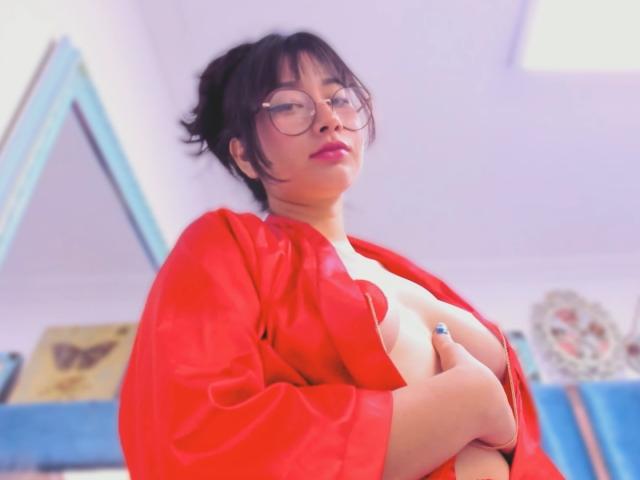 Genesiss, a medium-skinned woman with short black hair and glasses, stands with a confident expression. She wears a red, open robe that exposes her large breasts, which she covers with her hands. Her blue nail polish contrasts with her red robe. The image has a soft, slightly blurred effect, emphasizing her face and upper body. The background includes a blue-framed mirror and a floral decorative piece. The overall composition focuses on Genesiss's bold, self-assured presence.
