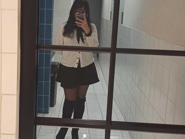 A mirror selfie of Genesiss in a bathroom. Genesiss has long, dark hair, wearing a white button-up shirt, black skirt, and black knee-high socks. Genesiss's right hand holds a phone, partially covering their face. The mirror has a black grid frame. The room has white tiled walls and floor, with a blue tiled section on the left. The reflection is slightly blurred. Genesiss's legs are positioned slightly apart, and the phone case has a dark pattern.