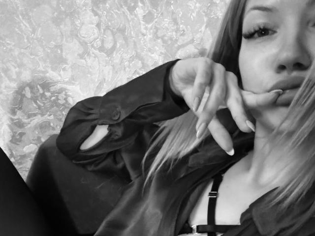Black-and-white selfie of XinitaBoom with long hair, pouty lips, and fingers near mouth. Wearing a dark shirt, visible bra, and textured wallpaper in background.