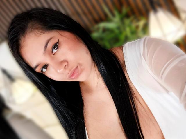 AnastasiaRouge has long black hair, brown eyes, and a pouty face with a slight nose up. She's wearing a white, open, sheer top, showing a bit of her chest.