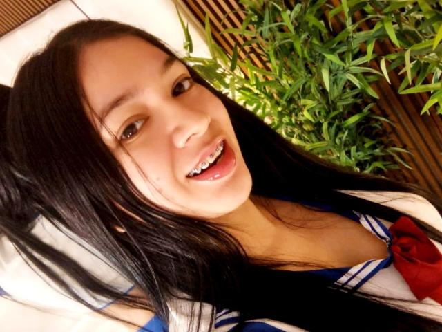 AnastasiaRouge, a young Asian woman with long black hair, is in a close-up selfie. She has smooth, light skin and a slight smile with her lips parted. She's wearing a blue and white sailor-style school uniform with a red bow. Her brown eyes are looking directly at the camera. The bamboo plant in the background adds greenery to the image. The lighting is warm, highlighting her facial features. The photo has a casual, intimate feel.