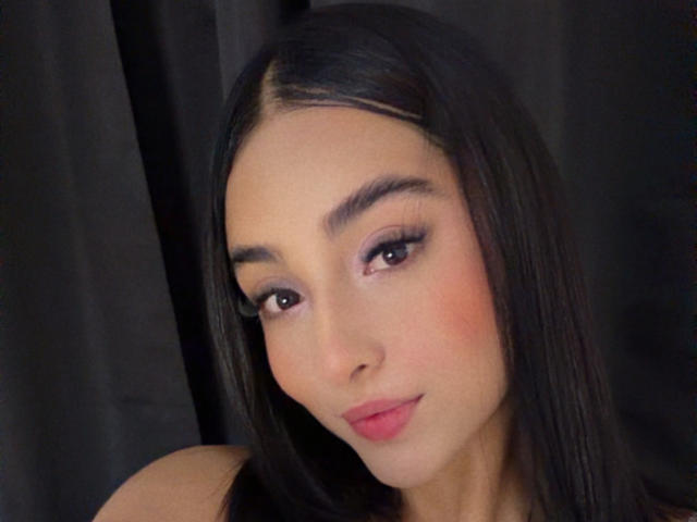 EmmaWalter has straight, dark brown hair parted in the middle. Her face is slightly angled to the right, with a subtle, confident smile. She has thick, dark eyebrows, long eyelashes, and pinkish eyelids with light purple eyeshadow. Her skin is smooth and light, with a slight blush on her cheeks. She has full, pink lips. The background is a dark, solid color, making her face the focal point. The image has a soft, warm light, highlighting her facial features. She is wearing a dark, strapless top.