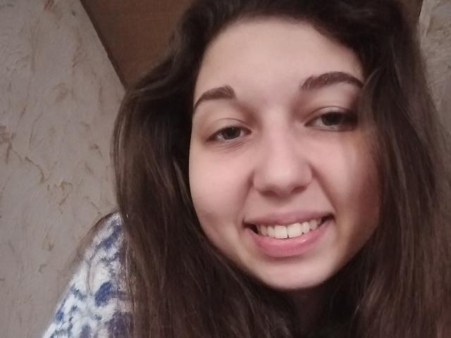 Close-up of Sanella with medium-length brown hair, slightly wavy, and dark eyebrows. She has a fair skin tone and is smiling with visible white teeth. She is wearing a blue and white patterned top. The image focuses on her face, which is centered in the frame. Her eyes are brown and slightly downcast. The lighting is even, highlighting her natural facial features. The texture of her hair is soft and slightly tousled.