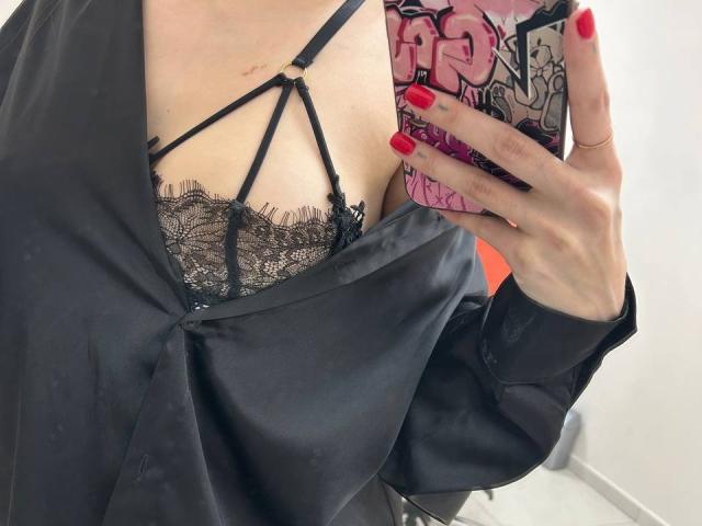 Close-up of MoonLiza's upper body. She's wearing a black strappy lace bra with intricate patterns, visible under a loose, black satin shirt. Her right hand, with red-painted nails, holds a phone with a pink and black graffiti-style case. The phone's case has bold, graffiti-style text and shapes. Her skin is light, and her fingers have a ring on the index finger. The image focuses on the contrast between the satin shirt and the detailed lace bra. The red nails and phone case add vibrant color to the otherwise monochromatic composition.