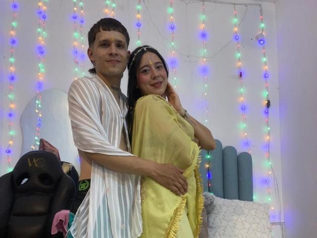 AnjaliRohan stands to the left, a young man with short, dark hair wearing a sheer, white robe. He has a medium build and is partially nude, with his hand on her hip. AnjaliRohan, a young woman with long, dark hair and a medium build, wears a yellow, semi-transparent shawl that covers her torso. She has a gentle smile and her right hand touches her hair. Multicolored, string lights hang in the background. The room has a blue, rolled-up mat and a black gaming chair with "LC" on it. The image has a casual, intimate feel.