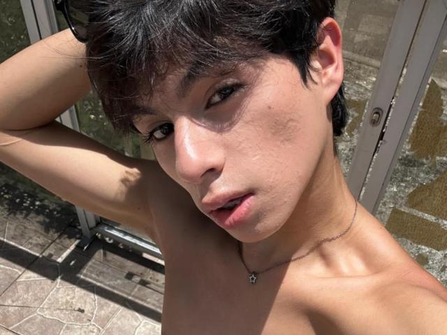 SaintDangelo has short black hair, fair skin with a slight tan, and visible acne on the left cheek. SaintDangelo's pink lips are slightly parted, and they're wearing a necklace.