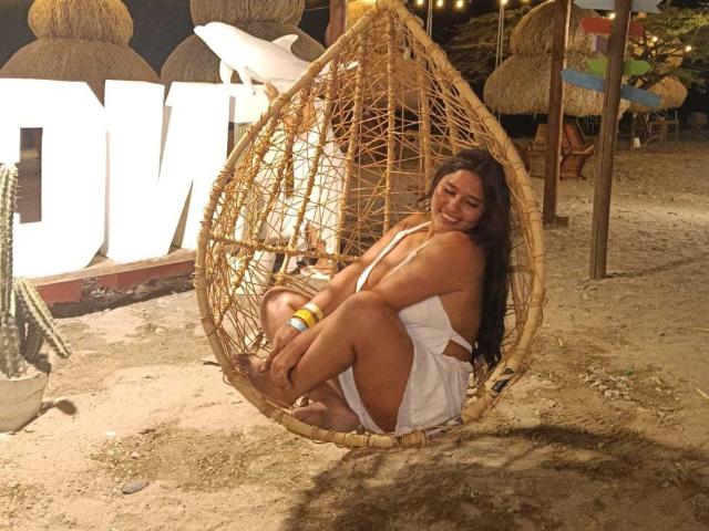GemmaBellini sits in a woven egg chair on a sandy ground. She has long black hair and a curvy, medium-build. She wears a white, backless dress and a yellow bracelet. Her legs are bent, and she's smiling at the camera. The chair's texture is coarse with visible weave patterns. The light is warm, and there's a cactus in the left background. The image has a casual, beachy vibe.