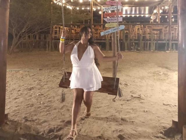 GemmaBellini swings on a wooden seat in a sandy beach area at night. She wears a white, sleeveless, deep V-neck dress, and sandals. Her dark hair is down, and she holds the ropes. String lights hang above, with signs like "Dance" and "Snack Bar" visible. Her right wrist has a yellow bracelet. The scene is warm-toned and slightly blurry.