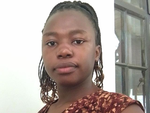 Yollah has dark brown skin, small nose, and thin lips with a slight mustache. She's wearing a brown, patterned top and has her hair in neat, medium-length twists.