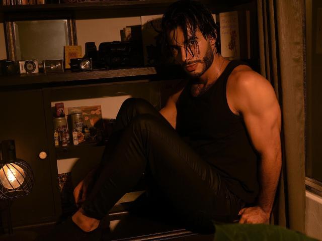 ValentinDurand sits on a shelf, wearing a black tank top and wet black pants, with muscular arms and wet, dark hair, looking at the camera with a serious expression.