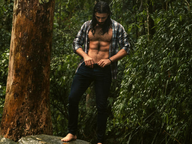 ValentinDurand with long black hair, shirt unbuttoned, exposing a muscular, bare chest, and black jeans, standing barefoot on a rock in a forest, hands near his jeans.