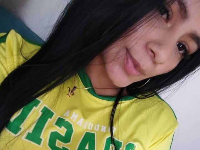 Close-up of SharickLion with long black hair, slightly smiling, wearing a yellow Brazilian soccer jersey with green trim and "AMERICA" in bold green letters. Small gold necklace with a pendant visible. SharickLion has light brown skin, small hoop earrings, and brown eyes. Slightly messy hair, soft light on face. Image has a casual, selfie-style composition.