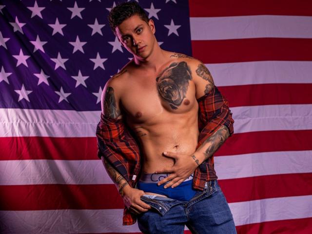 JeanBroc, shirtless, with tattoos, red plaid shirt off, blue jeans, hand on exposed blue underwear, standing in front of American flag, muscular, short brown hair, serious expression.
