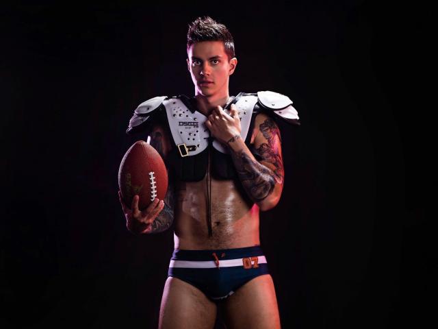 JeanBroc with tattoos, muscular, holding football, wearing black and white football armor, and blue briefs, standing against dark background, looking serious, short brown hair, slight facial stubble.
