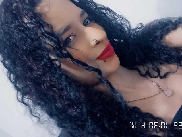 MeganKatty with curly black hair, nose ring, lip piercing, red lipstick, and necklace, looking at camera with a slight smile, showing her face and upper chest.