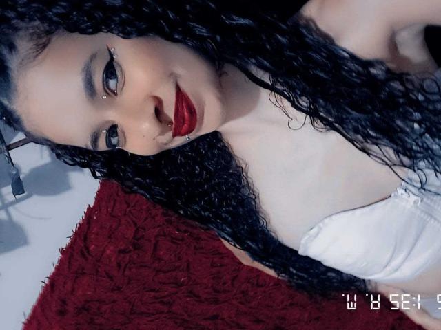 MeganKatty with curly black hair, red lipstick, and white bra, showing off her tits, pale skin, and nose ring, looking at the camera with a slight smile.