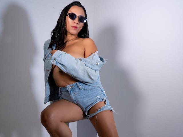 Jade Foster, avec ses cheveux foncés, portant des lunettes de soleil, une veste en jean bleu clair que porte négligemment sur ses épaules, et un short en jean bleu déchiré, laissant entrevoir ses seins et un petit tatouage sur son côté droit..