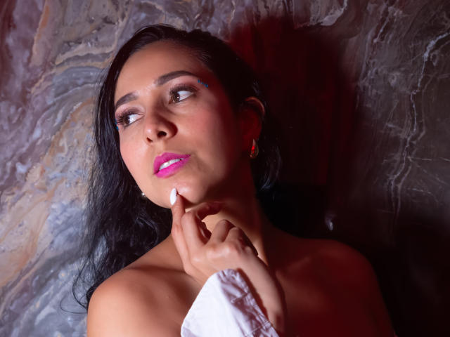 KattyRivera with pink lipstick, dark wet hair, and white nail, touching chin, looking up, red light on face, marble wall, earrings, bare shoulders, slight smile, sexy.