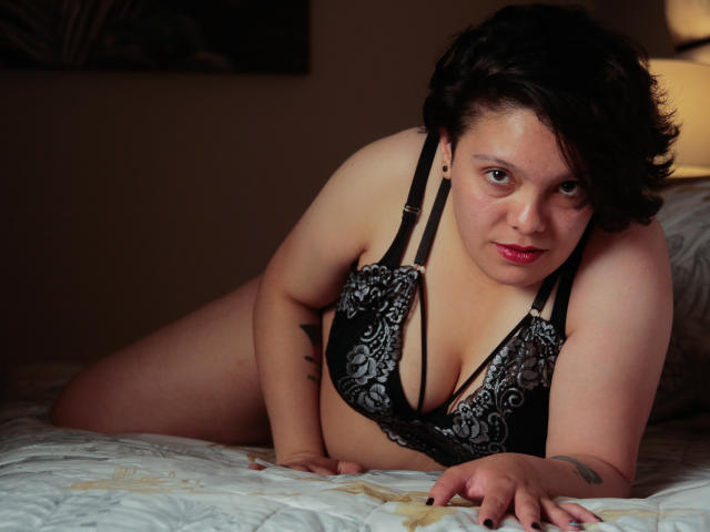 GabriielaStone, com cabelo preto curto, batom vermelho e um sutiã de renda preta, está na cama, inclinada para frente, mostrando seus seios e uma tatuagem no braço direito..