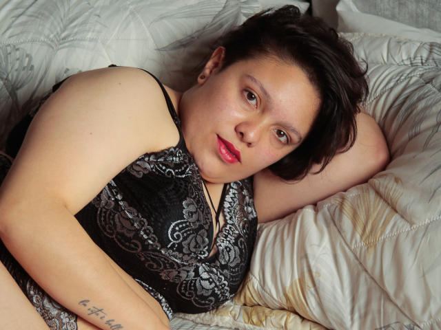 GabriielaStone, met kort zwart haar, rode lippenstift en een tatoeage op haar rechterarm, liggend op een wit bed, gekleed in een zwarte kanten top, kijkend naar de camera..