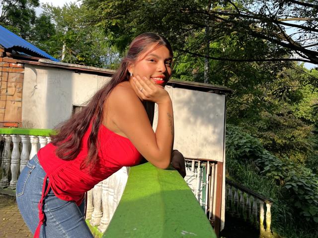 AlyssaGreen, com cabelos castanhos longos, um top vermelho sem alças e jeans azuis, apoiada em um corrimão verde, com a mão no queixo, sorrindo, mostrando seus seios e nádegas, sob a luz do sol..