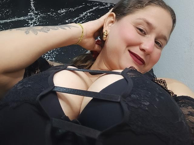 AlexaHotSeins with large, black-laced tits, black bra, and cleavage, smiling, dark lipstick, gold earrings, yellow bracelet, and tattoo on arm, in a selfie.