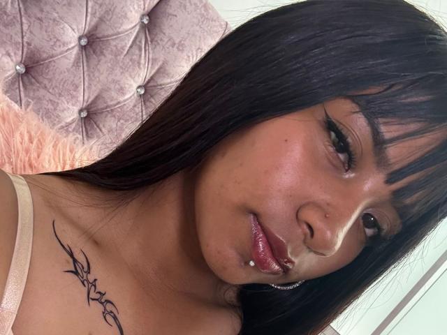 NahomyNoir has dark brown straight hair, a nose piercing, and a lip piercing. She has a tattoo on her left shoulder and is wearing a pinkish-beige top.