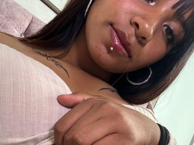 NahomyNoir's face, showing a nose ring, lip piercing, and ringed ear, with a hand on her tits, wearing a ribbed pink top, tattoos on her chest.