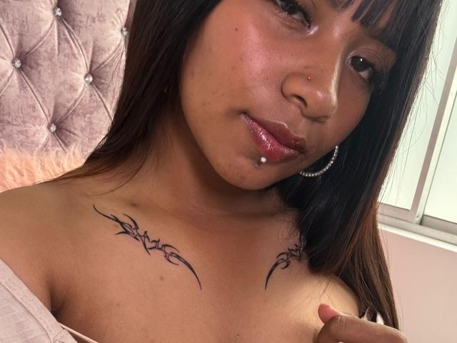 NahomyNoir has a tan skin tone, dark brown hair, and a nose ring. She's topless with tribal tattoos on her tits, a lip piercing, and silver hoop earrings.