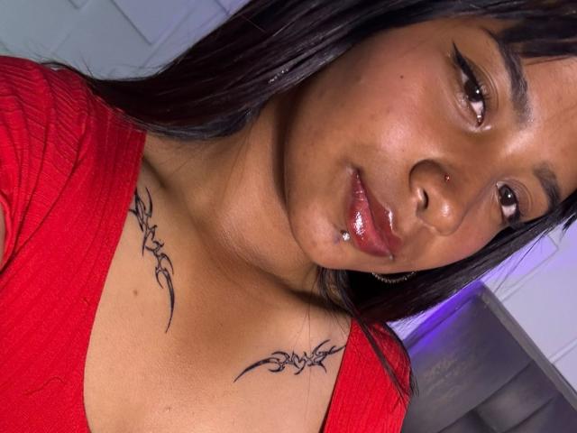 NahomyNoir with dark brown skin, glossy pink lips, black tattoo on chest, red top, and long black hair. Boobs visible, subtle nose ring. Soft light on face.