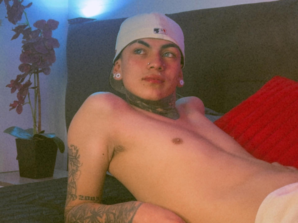 AdamsWalker is a shirtless dude with tattoos, wearing a white cap, and a nose ring, lying on a bed with a red pillow, looking up. His pants are down.