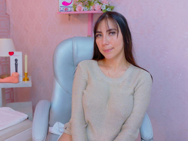 FreyaraVenwo with long brown hair, small tits, wearing a beige sweater, sitting in a light blue chair, pink wall in the background, small shelf with candles and toys.