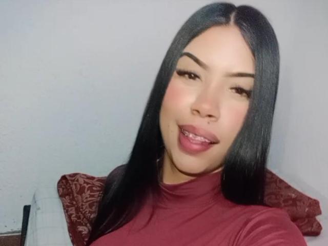 SharlotTeam has long black hair, a nose ring, and is smiling with pink lips, wearing a red turtleneck, showing her teeth. Her brown eyes are slightly winking.