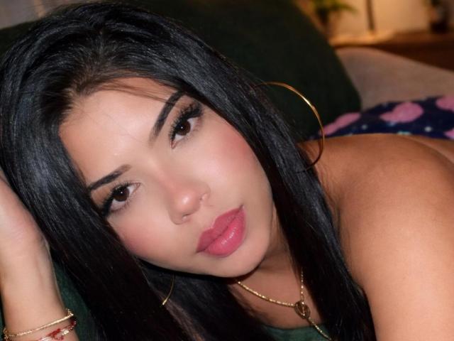 SharlotTeam with long black hair, hoop earrings, pink lipstick, and brown eyes, lying down with a pouty expression, wearing a gold necklace and bracelet, looking directly at camera.