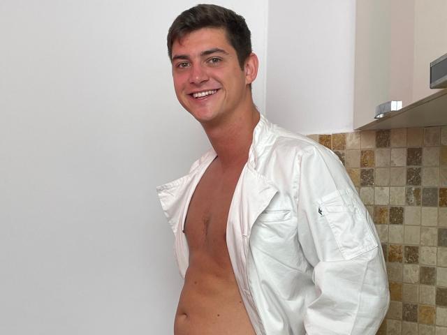 AndyFame with short brown hair, smiling, shirt open showing a toned, bare chest, white shirt, hands in pockets, standing in a kitchen with tiled wall.