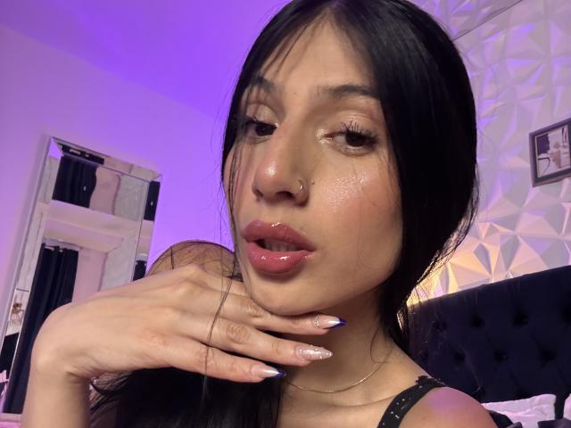 SophieBeaumont has shiny skin, glossy pink lips, and long black nails, with her hand on her chin, wearing a nose ring, and looking seductive with purple lighting.